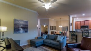 A living room with a sectional sofa, a coffee table, and a TV. There is a kitchen area with a microwave and a bar. - Available 2025 !! Beautiful Water View 2 Bed+ Den +Garage Veranda Treviso Bay (Naples)