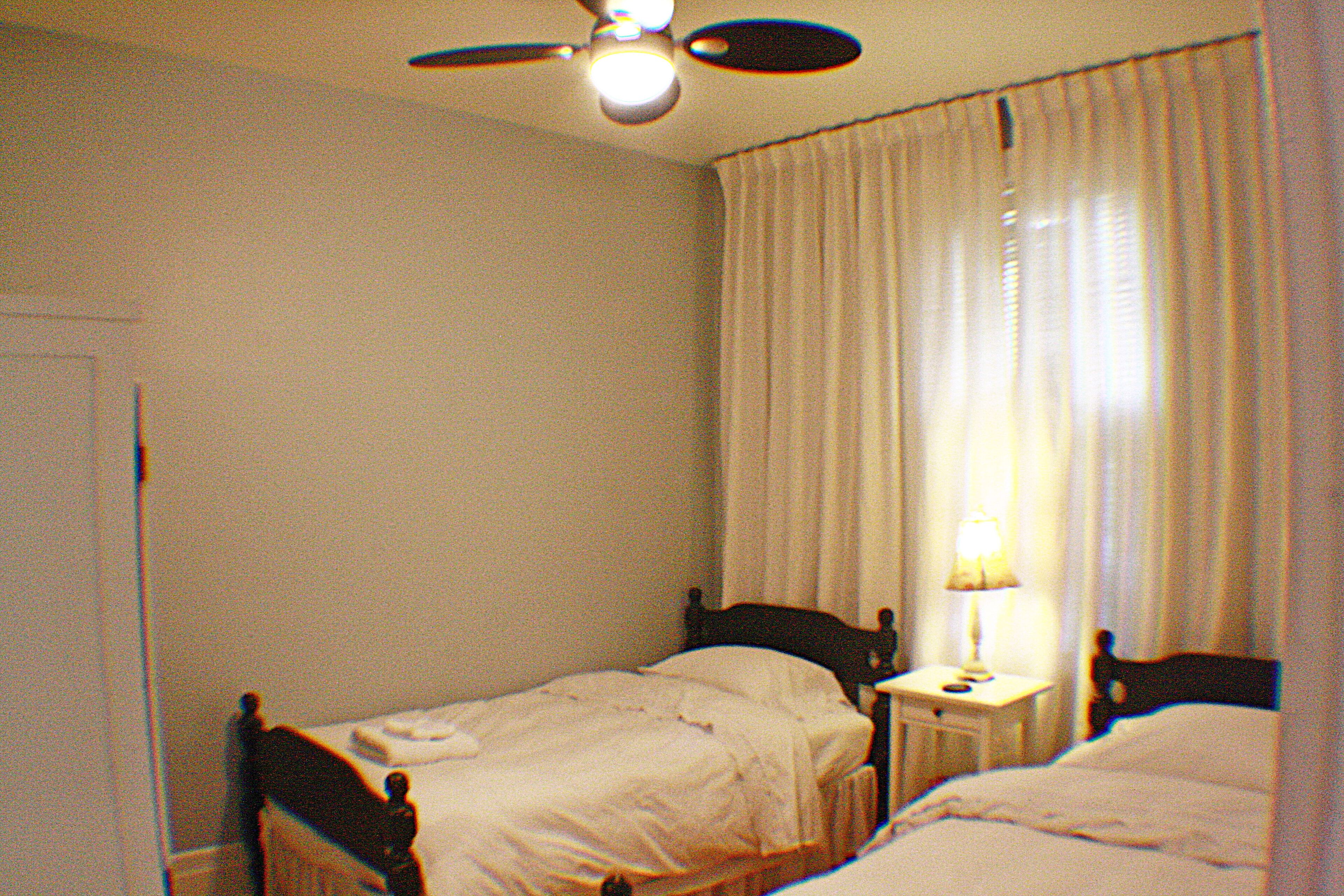 Une petite chambre avec un lit simple, un ventilateur de plafond, une table de chevet avec une lampe et une fenêtre avec des rideaux.