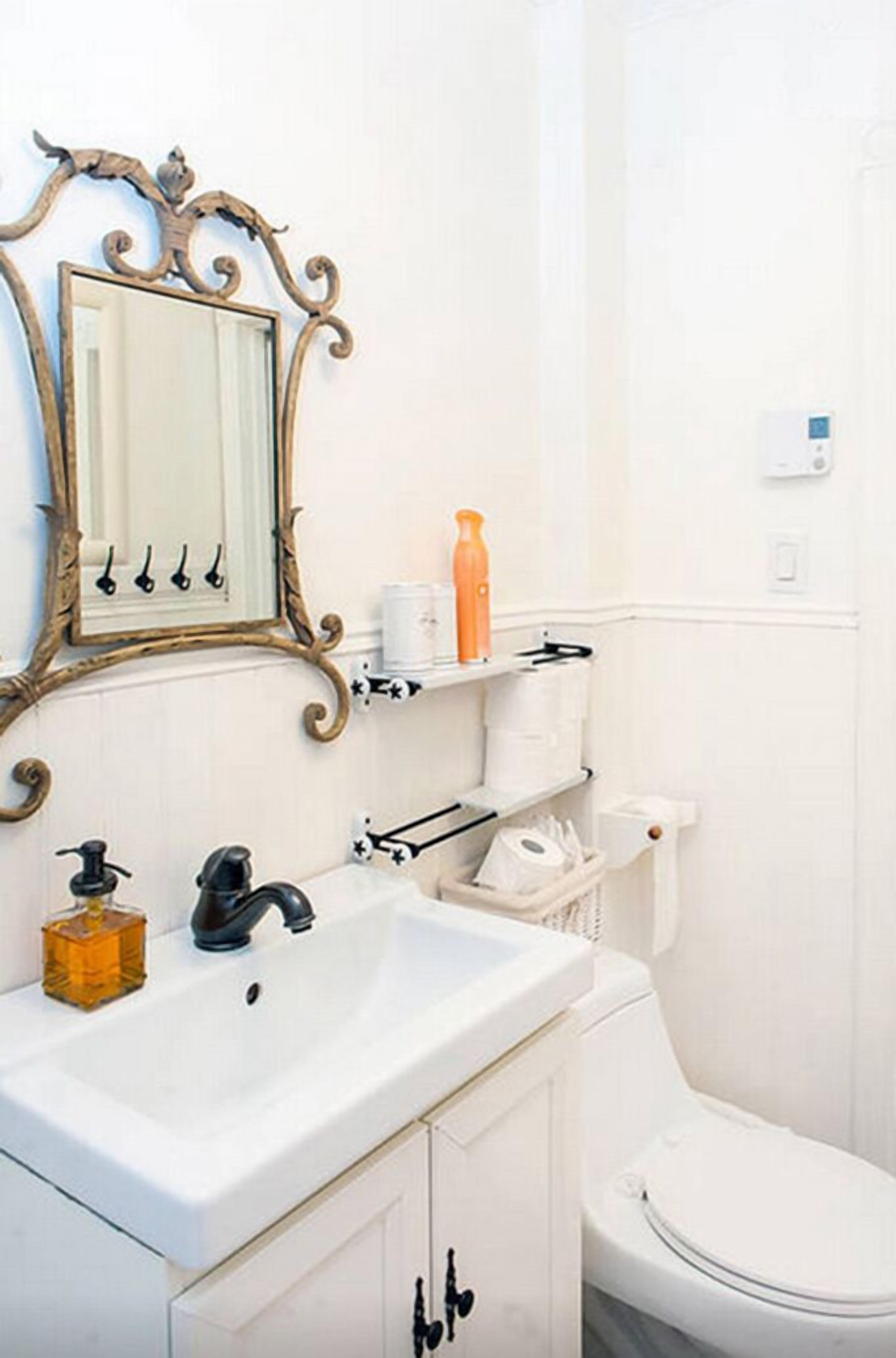 Un baño con lavamanos blanco, un espejo con marco decorativo, un inodoro y estantes con artículos de aseo.