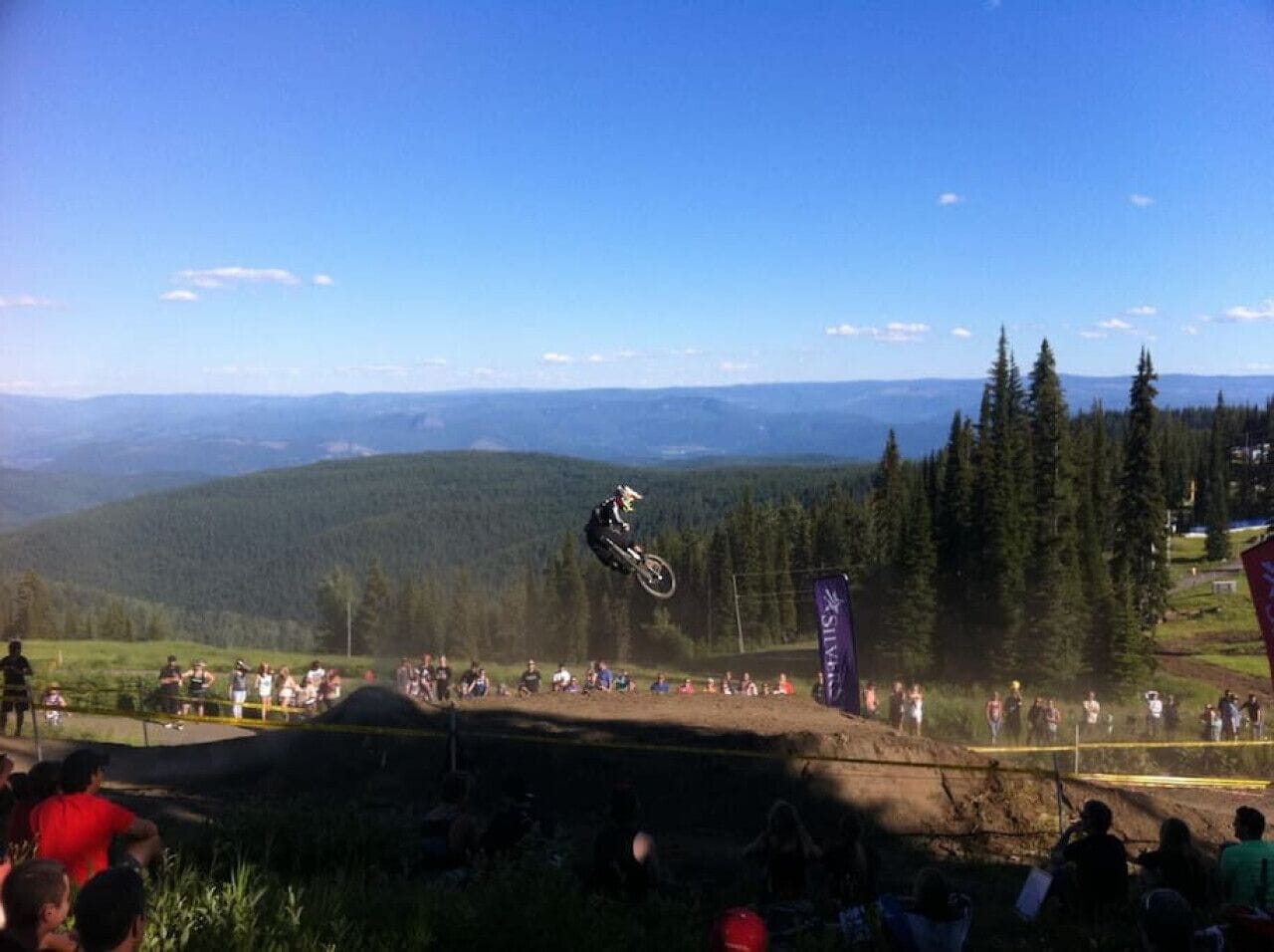 A motocross event with a rider performing a jump over a dirt track, surrounded by spectators and a mountainous backdrop.