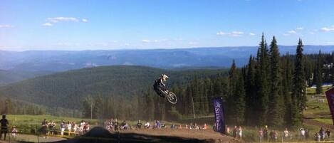 A motocross event with a rider performing a jump over a dirt track, surrounded by spectators and a mountainous backdrop.
