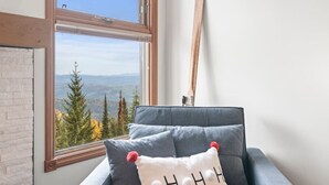 Quarto com vista para uma paisagem montanhosa através de uma janela ampla, piso de madeira e um sofá cinza com almofada branca.