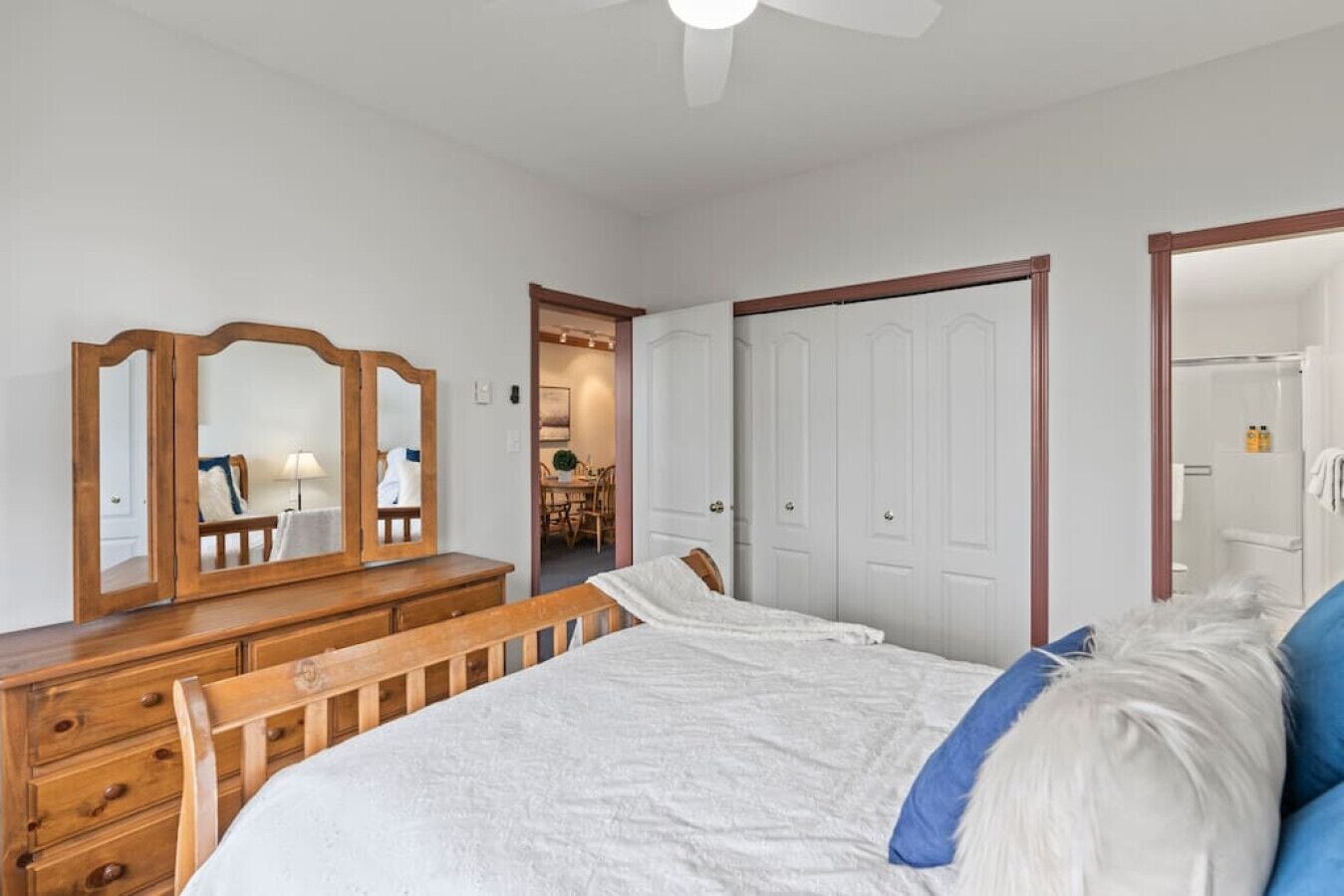 Un dormitorio con un aparador de madera, un espejo grande, una cama con ropa de cama blanca y una almohada azul.