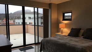 Ein Hotelzimmer mit einer großen Glasschiebetür, die auf einen Balkon führt, einem Bett mit grauer Bettdecke, einem Nachttisch mit Lampe und einem gerahmten Bild an der Wand.