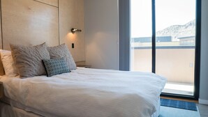 Ein ordentlich bezogenes Bett mit weißer Decke und mehreren Kissen, ein Holzkopfteil, eine an der Wand befestigte Lampe und ein großes Fenster mit Blick auf Gebäude und Hügel.