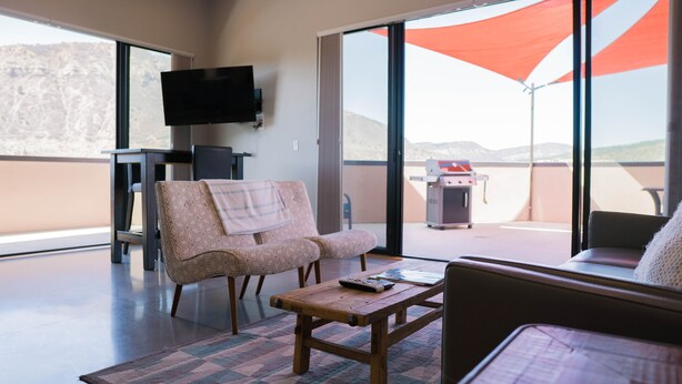 Ein Zimmer mit einer Couch, einem Sessel, einem kleinen Tisch, einem an der Wand befestigten Fernseher und Blick ins Freie durch große Schiebetüren.