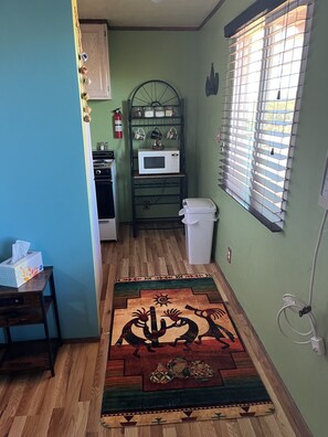 Una pequeña cocina con microondas, estufa y lavamanos. Hay una alfombra estampada en el suelo y una ventana con persianas.