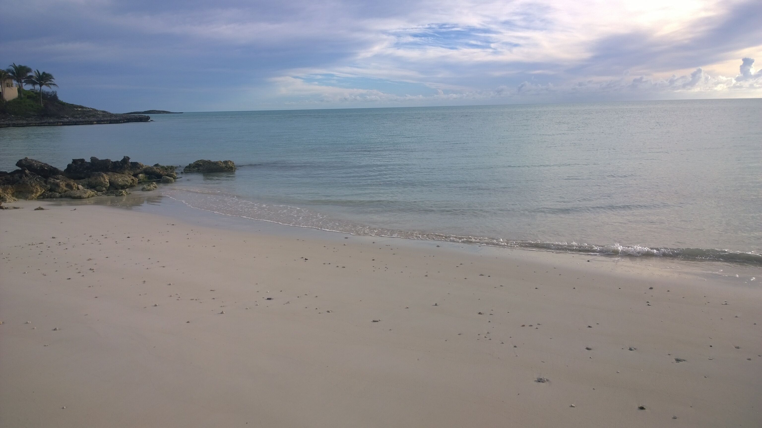 A beach with a calm sea, a sandy shore, and a rocky outcrop with vegetation.