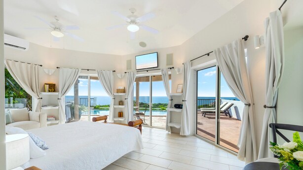 A spacious bedroom with a large bed, a ceiling fan, a TV, and a balcony with a view of the ocean.