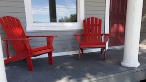 Twee rode Adirondack-stoelen op een veranda met een witte deur en een raam dat uitkijkt op een wateroppervlak.