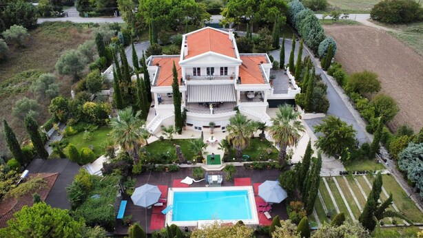 A large, white, two-story house with a red-tiled roof, surrounded by lush greenery and a swimming pool. - Villa Elena Perea Thessaloniki (Perea Thessaloniki)
