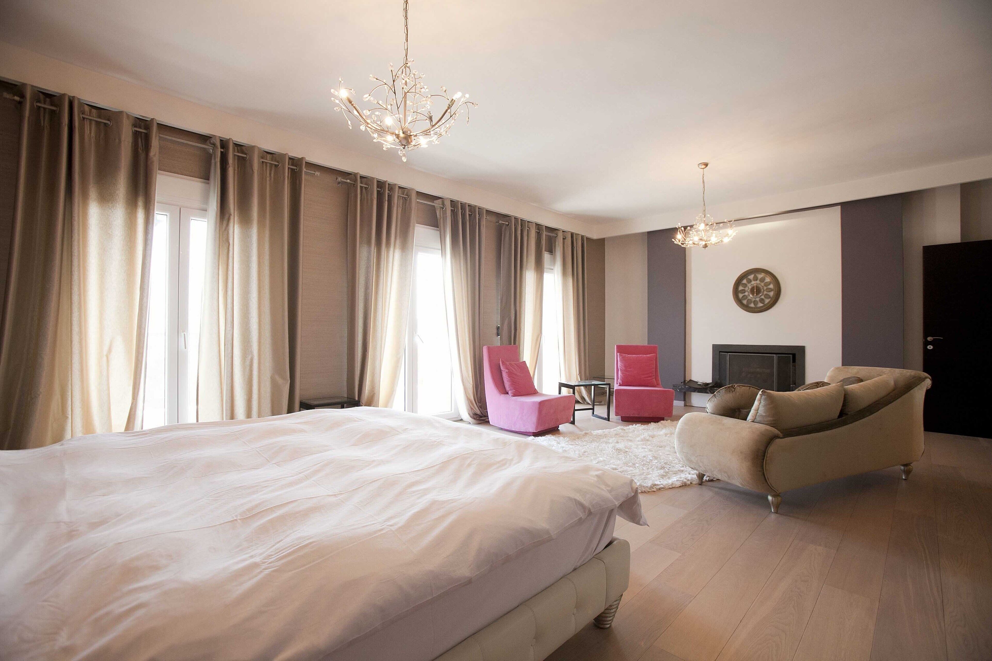 A bedroom with a large bed, two chairs, a sofa, a chandelier, and a fireplace.