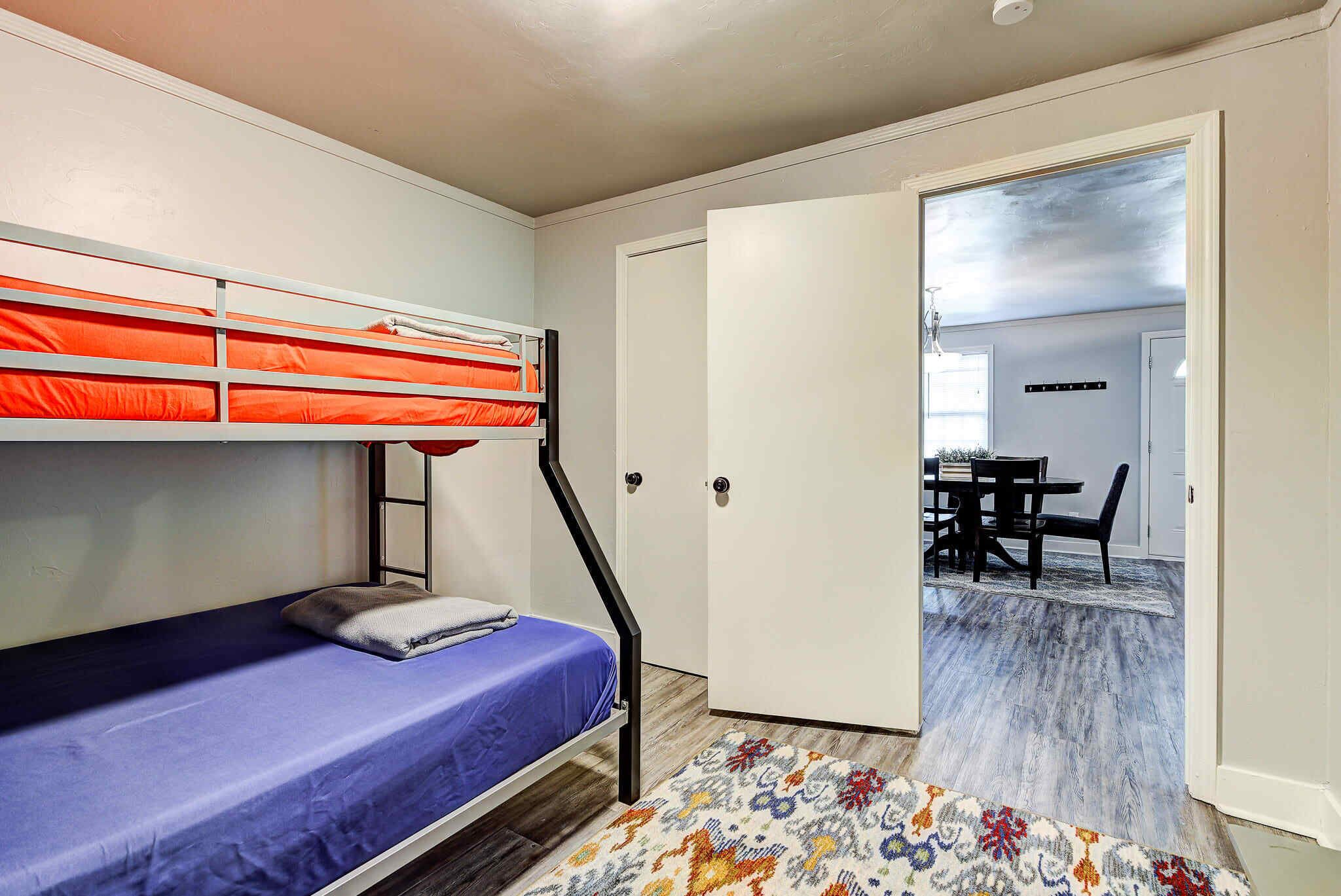 A bunk bed with a blue mattress, a white door, and a view into a dining area with a table and chairs.