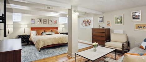 Un dormitorio bien iluminado con cama, mesitas de noche, cómoda y mesita auxiliar. Cuenta con una sala de estar con sofá, mesa de centro y sillón.