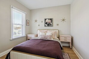 A bedroom with a bed, two bedside tables, a window with blinds, and wall decorations.