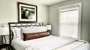 A neatly made bed with white bedding and a brown accent pillow, a framed artwork above the bed, and a window with blinds to the right. - Modern Farmhouse near VMI, W&L, SVU & VA Horse Ctr (Lexington)