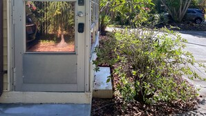 Une maison avec une porte donnant sur un porche abrité, entourée d’une végétation luxuriante et avec une voiture garée à proximité.