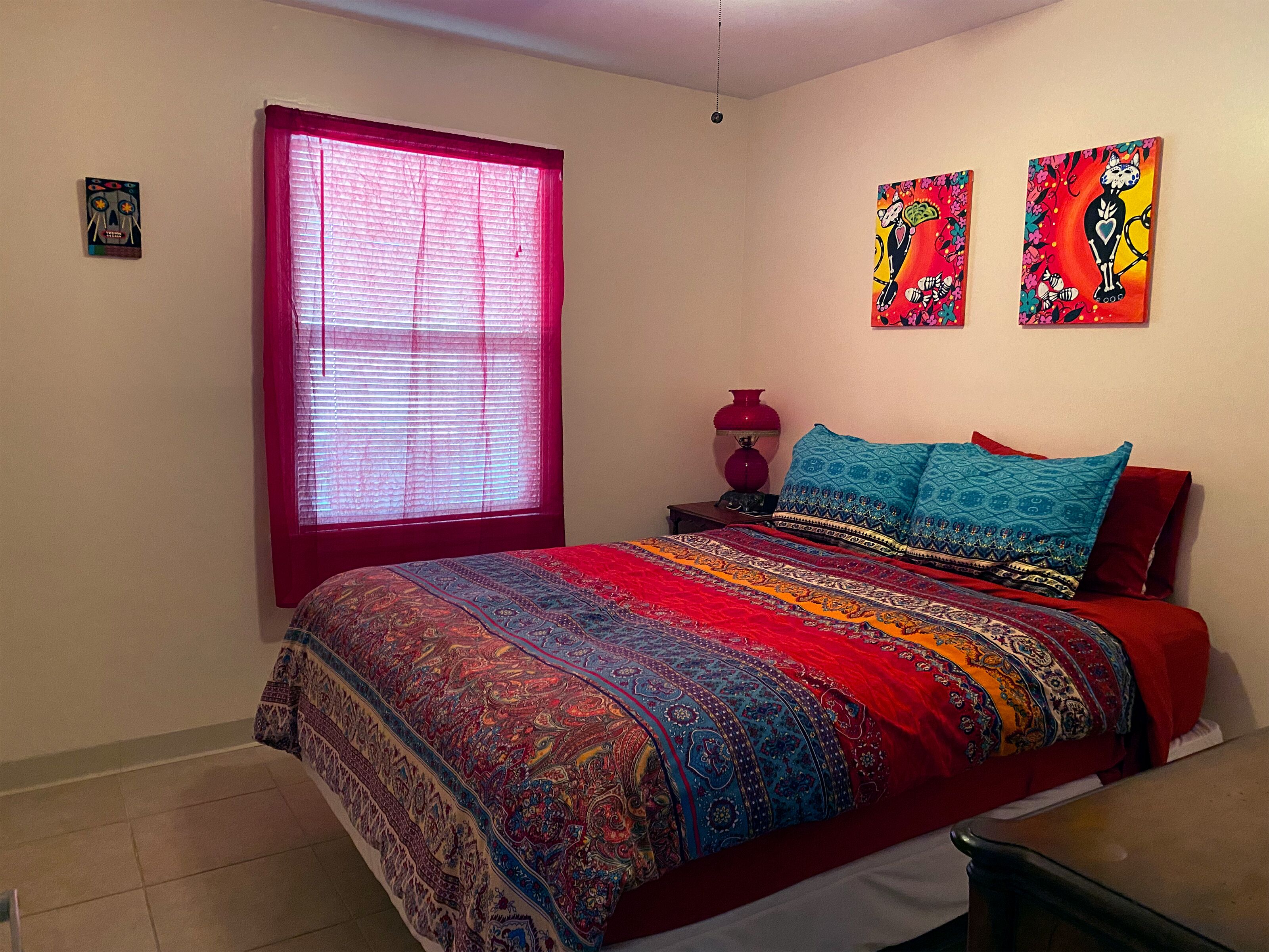 A bedroom with a large bed, a window with red blinds, a nightstand with a lamp, and two wall art pieces.