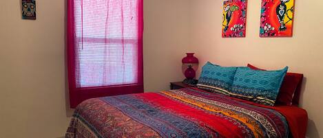 A bedroom with a large bed, a window with red blinds, a nightstand with a lamp, and two wall art pieces.