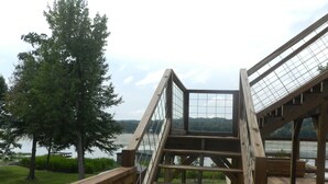 Eine Holztreppe mit Metallgeländer führt zu einer Terrasse mit Blick auf ein Gewässer, umgeben von viel Grün.