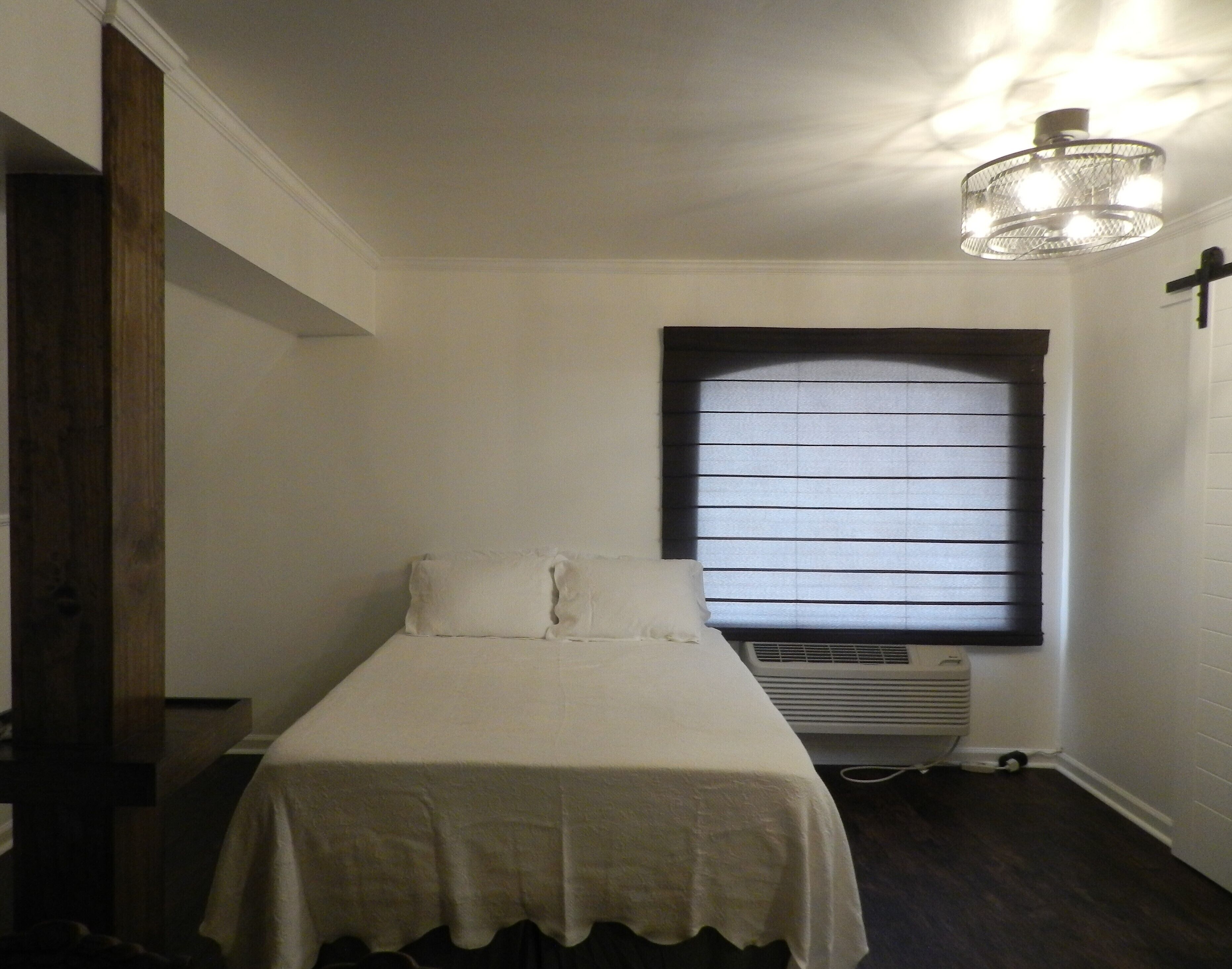 A small, neatly arranged bedroom with a single bed, a window with blinds, a ceiling light fixture, and a wall-mounted air conditioning unit.