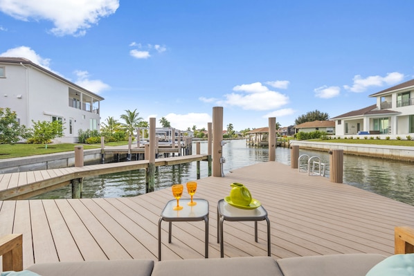 A waterfront deck with two tables, one with two glasses and a pitcher, and another with a single glass.
