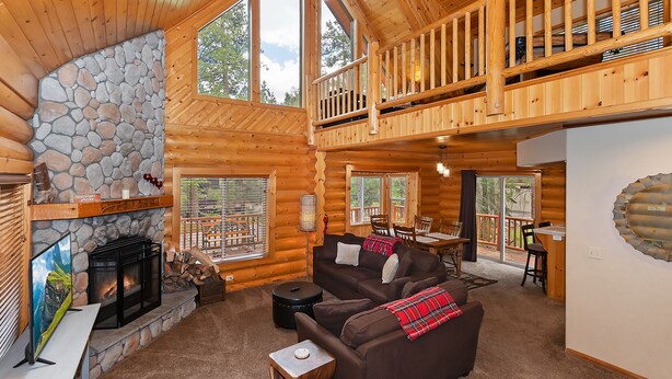 A spacious log cabin with a stone fireplace, a cozy living area with a sofa and chairs, a dining area with a table and chairs, and a balcony with wooden railings.