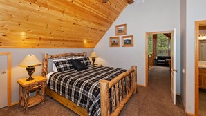 A cozy bedroom with a wooden ceiling, a bed with a plaid bedspread, two bedside lamps, and framed pictures on the wall.