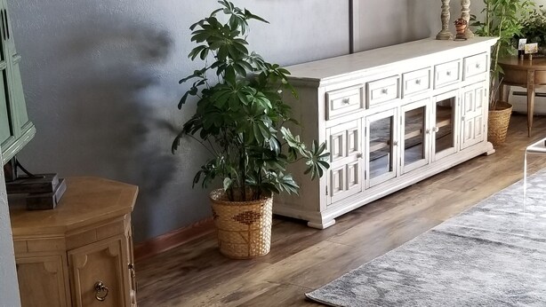 Una sala de estar con piso de madera, un mueble blanco, un televisor de pantalla plana en la pared y dos plantas en macetas.