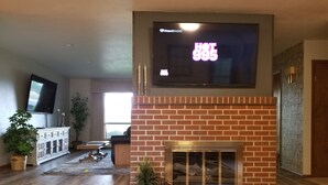Un salón con chimenea de ladrillo, un televisor montado que muestra 'HOT 99.5', y un mueble blanco con puertas de cristal.