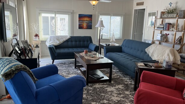 Una sala de estar con un sofá azul oscuro, un sillón rojo y otro sillón azul. Hay una mesa de centro con una lámpara, una estantería y un cuadro en la pared.