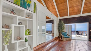 A spacious living area with a wooden ceiling, white shelves with decorative items, a wicker chair with a cushion, and a view of the beach through large sliding doors. - AS SEEN ON HGTV BEACH HUNTERS! Oceanfront, Amazing Whale Watching and Snorkeling (Captain Cook)