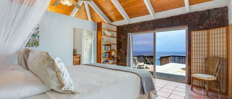 Une chambre avec un grand lit, un ventilateur de plafond, une bibliothèque et une vue sur l’océan.