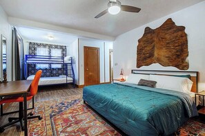A room with a bunk bed, a desk, a chair, a patterned rug, a bed with a blue bedspread, a wall-mounted headboard, a ceiling fan, and a door. - Eccentric Escape-hop, skip, jump to Plaza District (Oklahoma City)