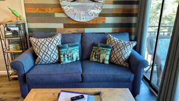 A cozy living room with a blue sofa, patterned cushions, a wooden coffee table with a remote control, and a decorative clock on the wall.