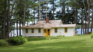 A white house with yellow trim and a chimney, surrounded by a grassy yard and tall trees.