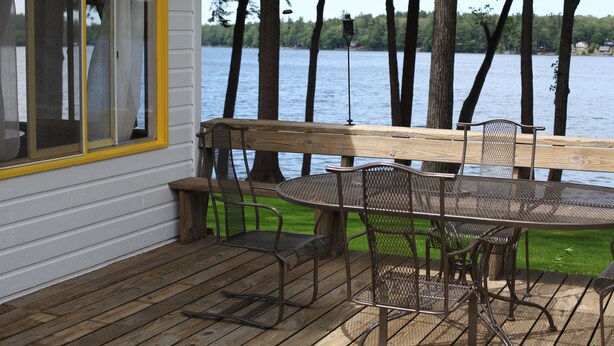 A wooden deck with a table and chairs, overlooking a lake with trees in the background.