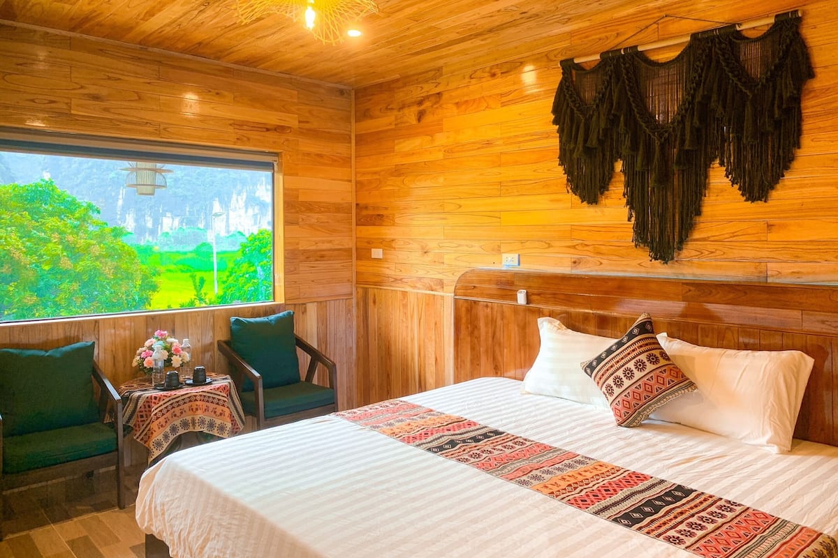 A wooden room with a bed, two chairs, a table with flowers, and a view of greenery outside.
