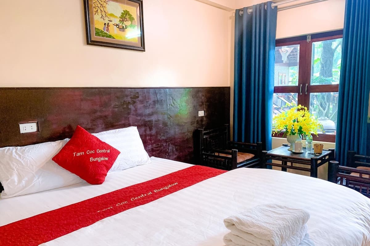 A neatly made bed with a red and white bedspread, a red pillow, and a white pillow. There is a framed picture on the wall above the bed, a small table with a vase of flowers, and a window with blue curtains.