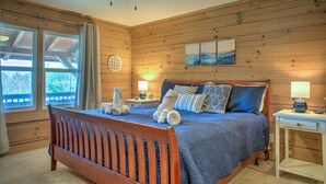 A bedroom with a wooden floor, a bed with blue bedding, a ceiling fan, and a window with curtains. - Pet-Friendly Log Cabin with Hot Tub & Biltmore Pass | 35 Min to Asheville (Waynesville)