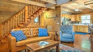 A wooden interior with a staircase, a leather sofa, a tufted armchair, and a kitchen area. - Pet-Friendly Log Cabin with Hot Tub & Biltmore Pass | 35 Min to Asheville (Waynesville)