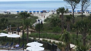 Un complejo turístico en la playa con tumbonas y sombrillas, un paseo marítimo que conduce a la arena, y palmeras.