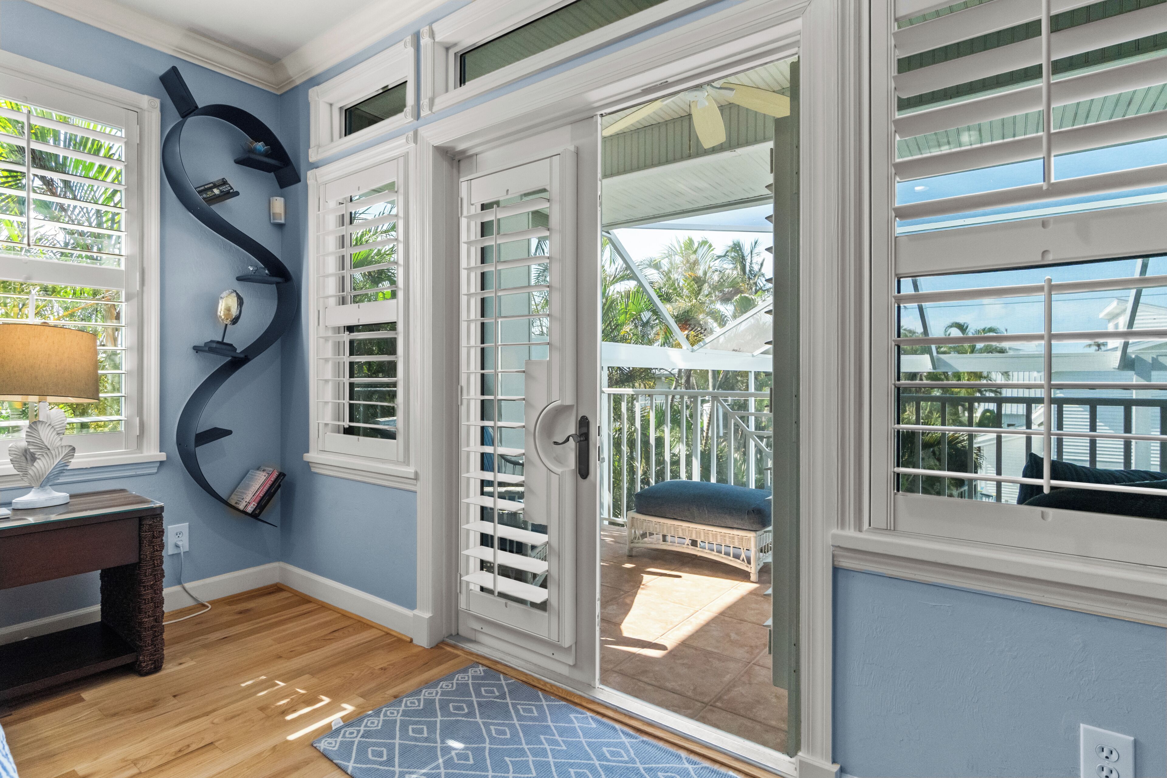Une pièce avec un plancher en bois, un bureau avec une lampe, un tapis bleu à motifs et une vue sur un balcon avec une chaise.