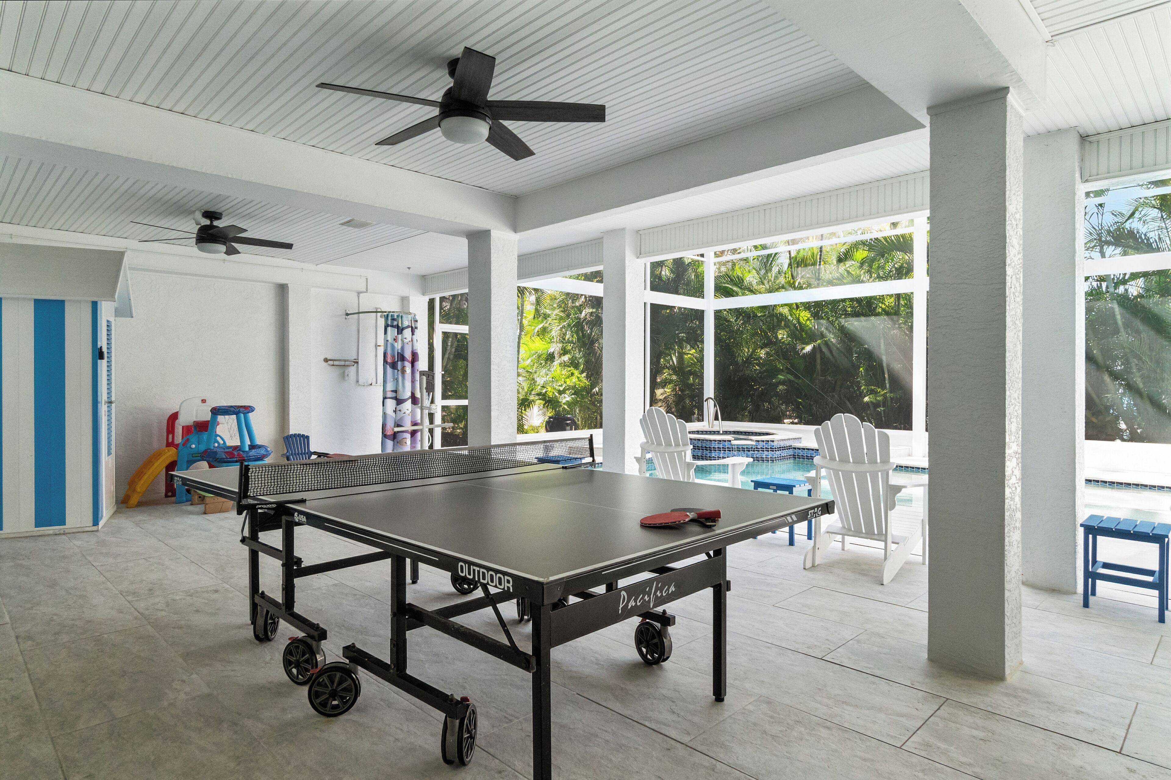 A ping pong table in a spacious room with large windows, ceiling fans, and a view of a swimming pool.