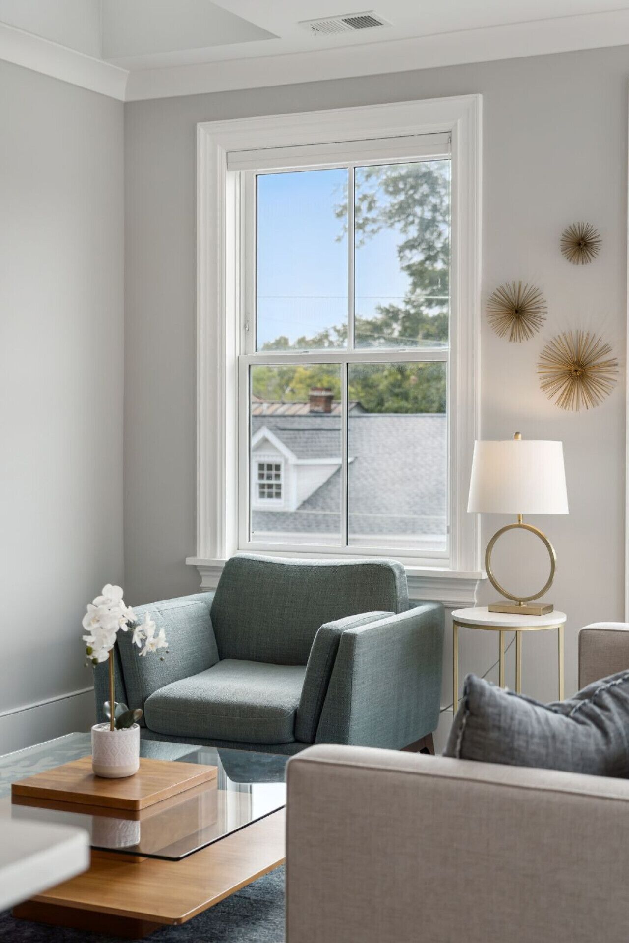 A living room with a grey couch, a side table with a lamp, and a window with a view of a house and trees.