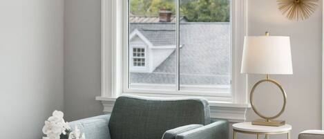 A living room with a grey couch, a side table with a lamp, and a window with a view of a house and trees.