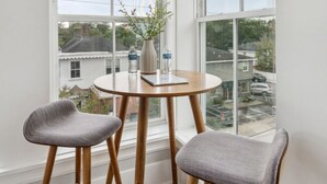 Habitación de esquina con una mesa redonda de madera y dos sillas grises tapizadas. En la mesa, un florero con plantas y botellas de agua.