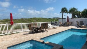 A swimming pool with a small hot tub, surrounded by lounge chairs and umbrellas, with a white fence and palm trees in the background. - Relaxed Cancellation Policy! Oceanfront home with pool,hot tub, fire pit, bikes. (St Augustine)