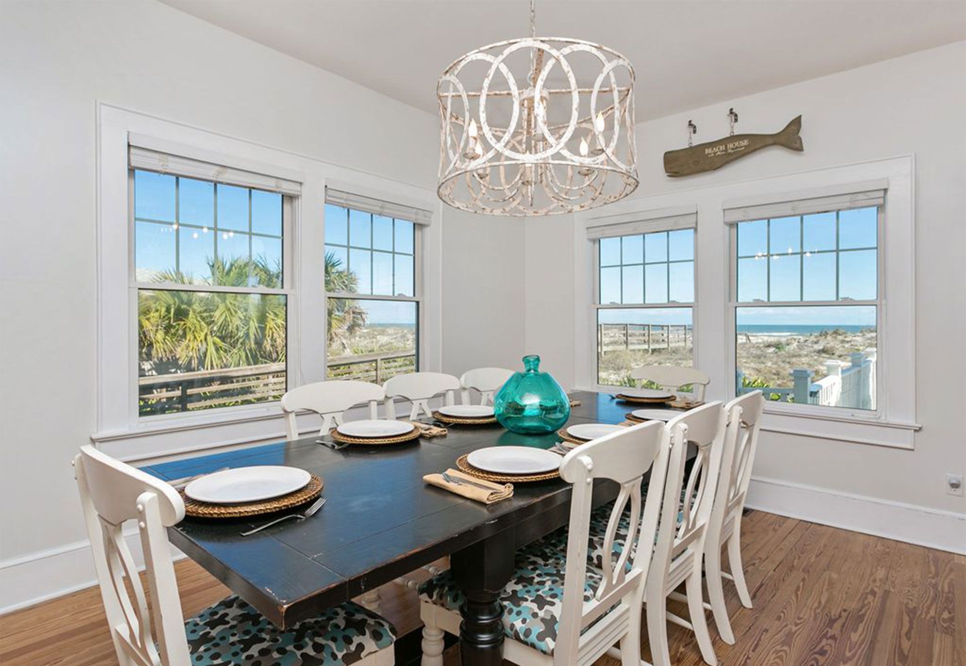 A dining room with a dark wooden table, white chairs with patterned cushions, and a view of the beach through large windows.