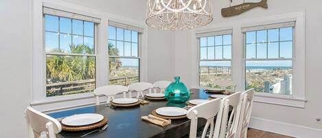 A dining room with a dark wooden table, white chairs with patterned cushions, and a view of the beach through large windows.